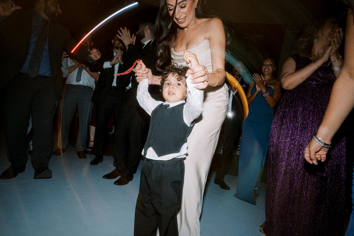 A young boy in a formal vest and tie dances joyfully at a lively wedding reception, surrounded by elegantly dressed guests