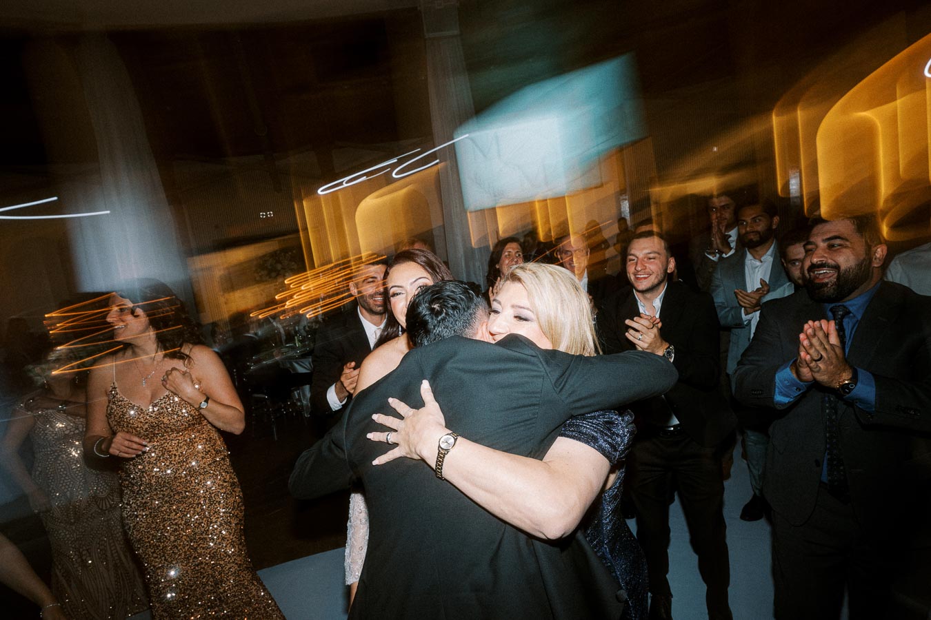 A joyful wedding celebration with guests hugging and clapping on the dance floor, featuring elegant attire and blurred