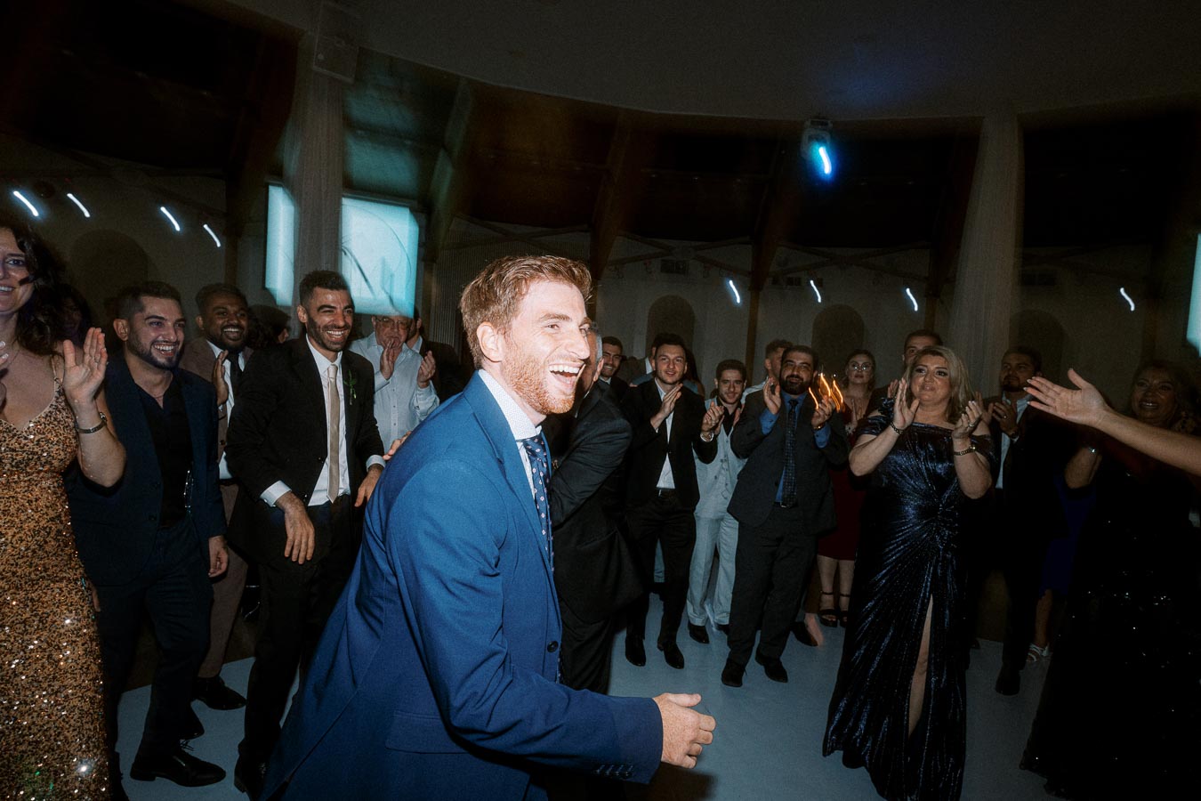 A joyful group of elegantly dressed people, smiling and clapping, gathered at a formal event in a dimly lit venue.