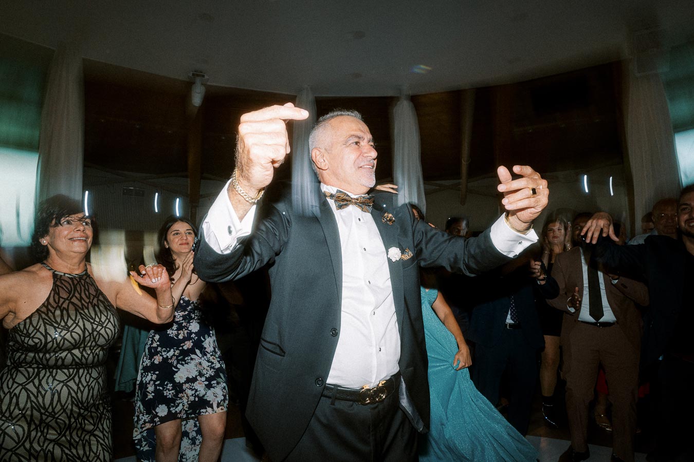 Senior man joyfully dancing in a suit at a lively wedding reception, surrounded by smiling guests in elegant attire.