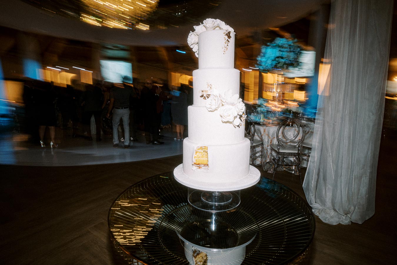 Elegant four-tier white wedding cake adorned with delicate floral decorations, displayed on a reflective table in
