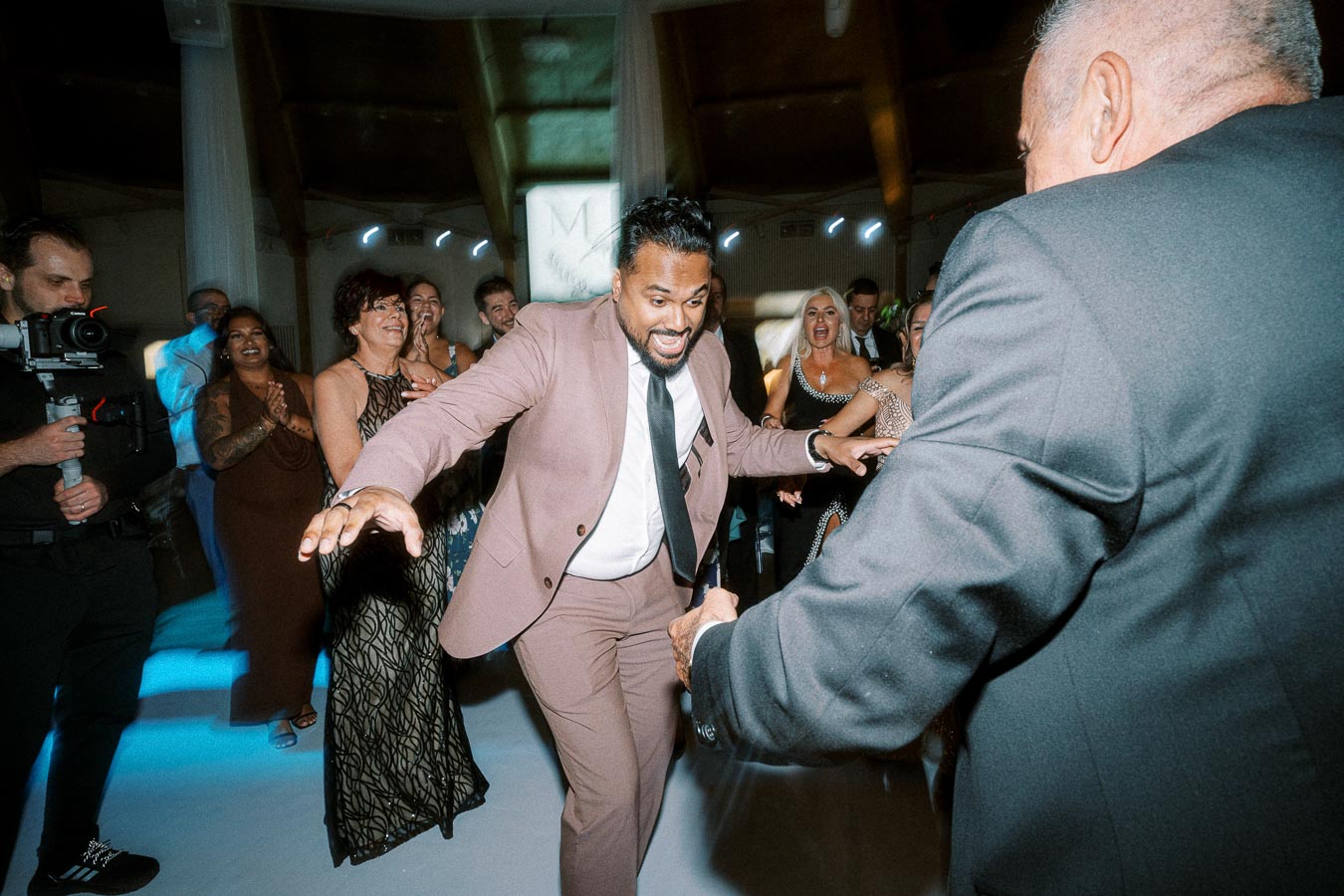 A group of people joyfully dancing and celebrating at a formal event, with a man in a pink suit engaging with the crowd,