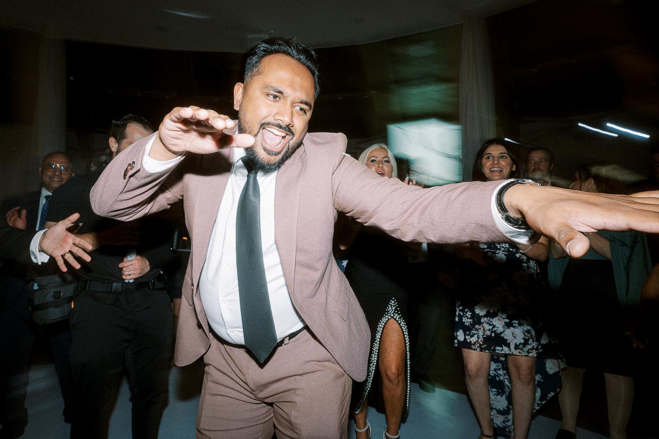 A man in a suit energetically dancing at an event, surrounded by smiling people in formal attire, creating a lively and
