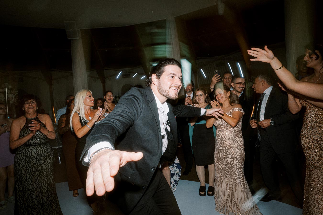 A joyful man in a suit dancing at a lively indoor party with a crowd of smiling and clapping guests.