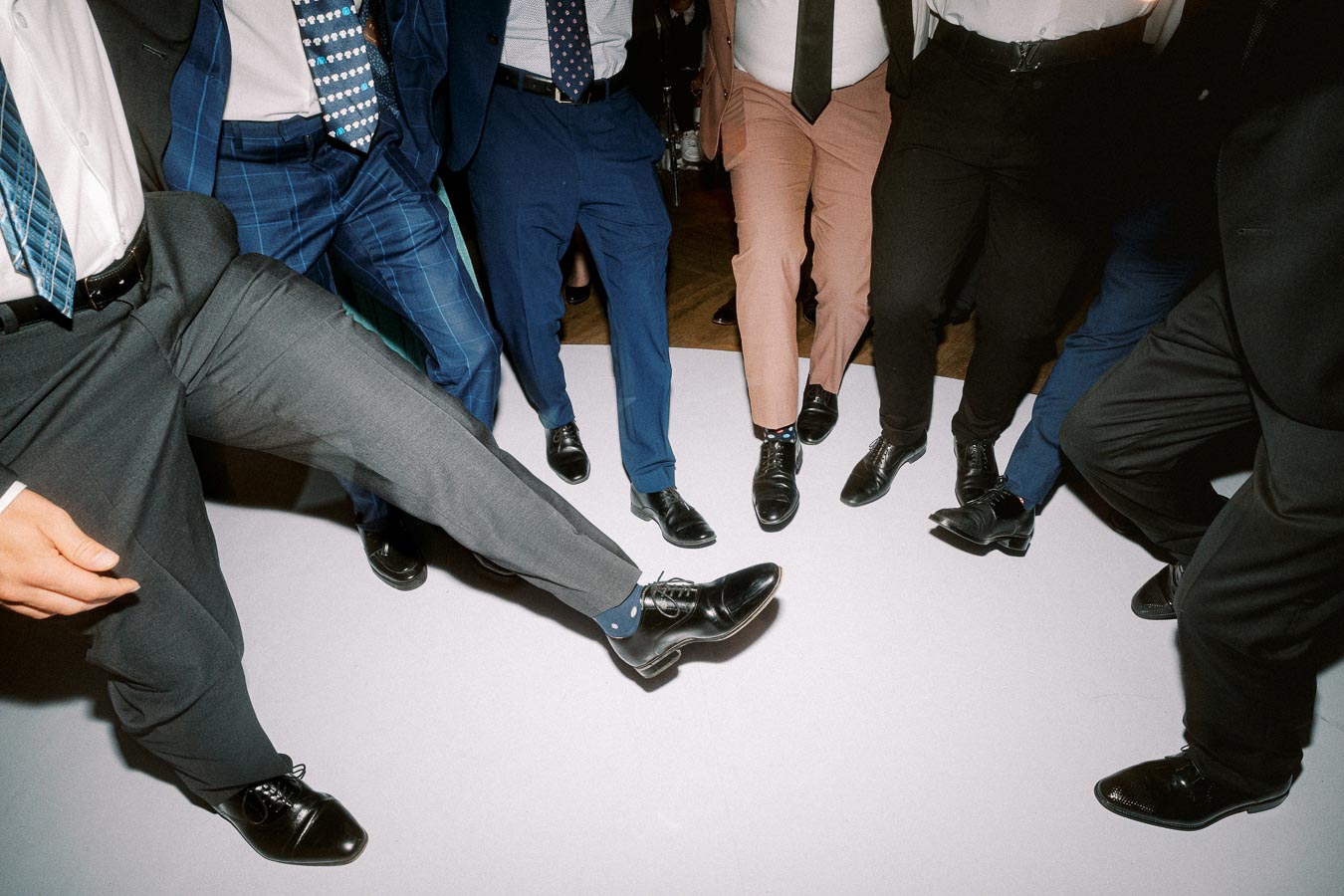 Group of men dressed in formal suits and polished shoes, playfully kicking their legs in a circle on a dance floor,