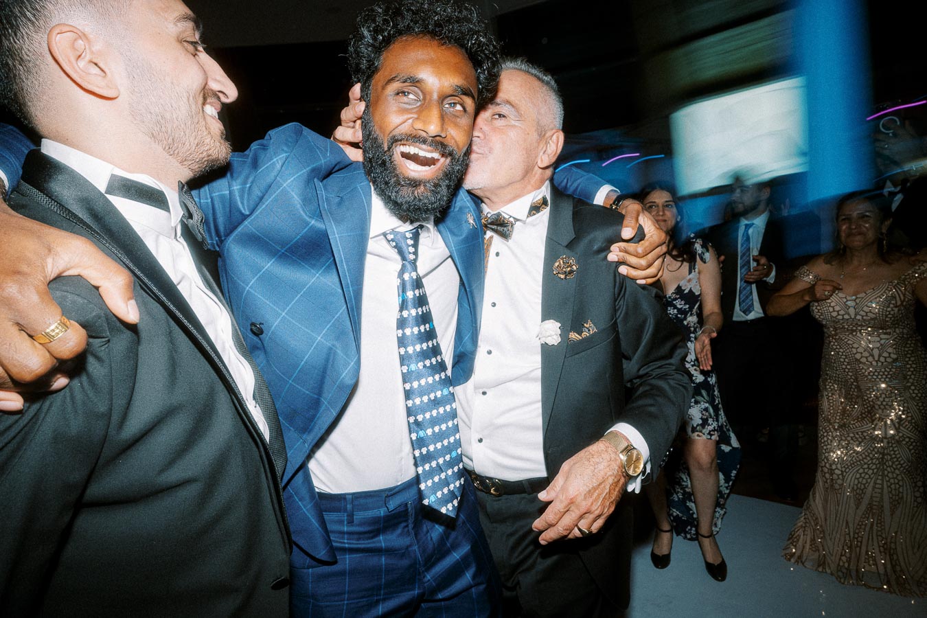 A group of well-dressed men laughing and celebrating at a lively party, surrounded by smiling guests and vibrant lighting,