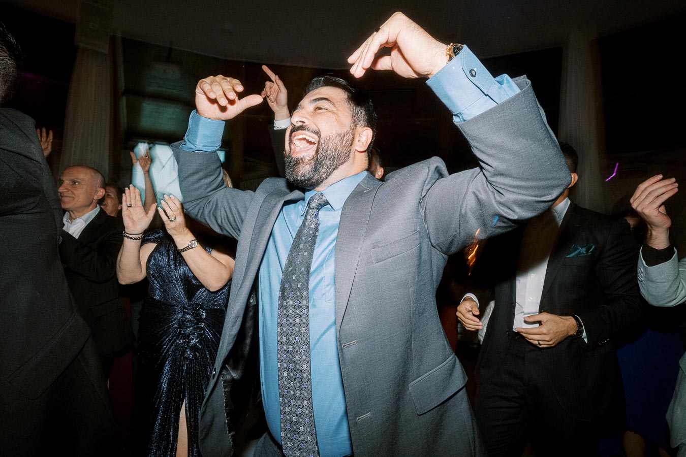 A joyful man in a suit enthusiastically dancing at a party with a crowd of people, featuring celebratory expressions and