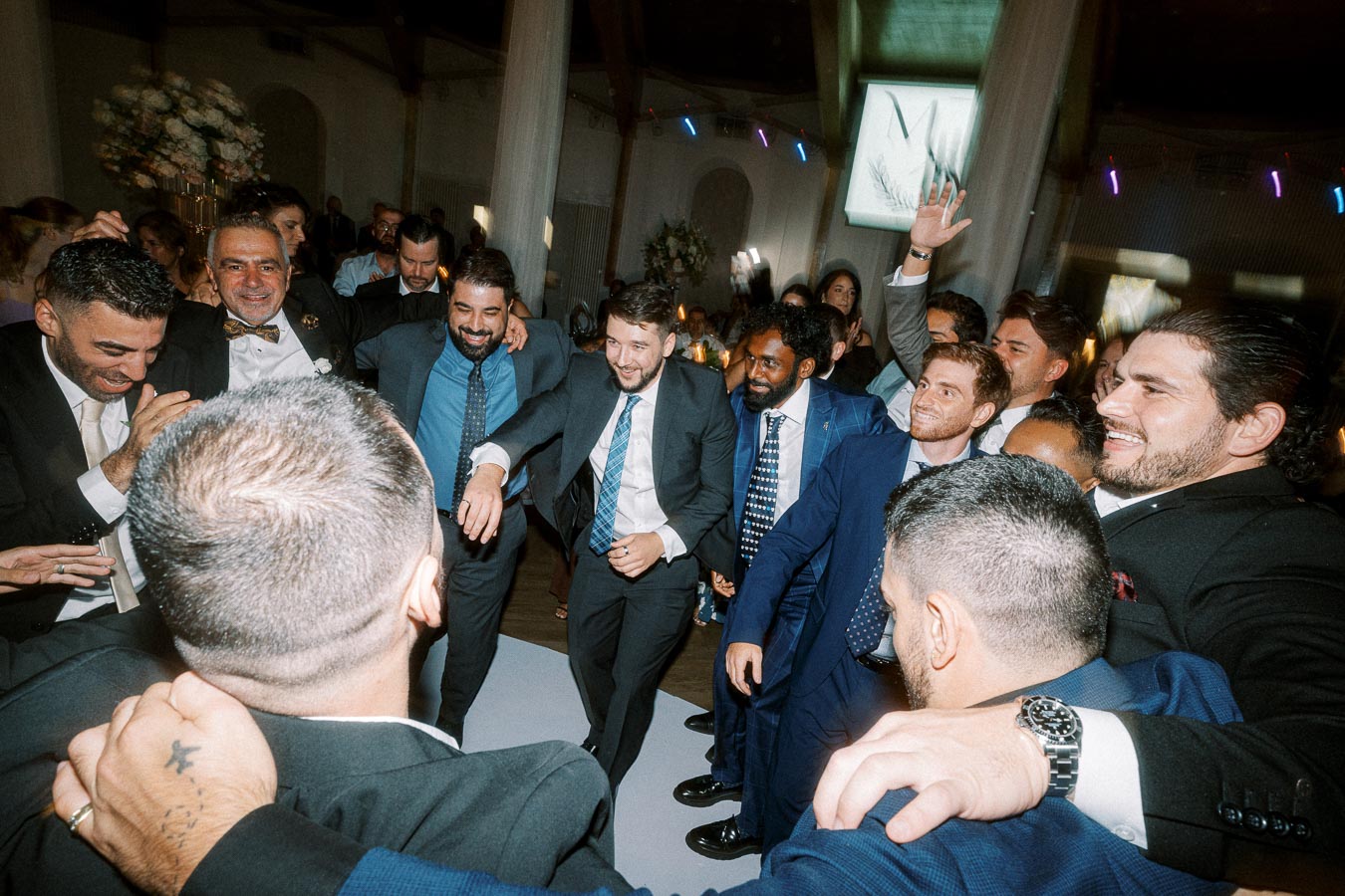 A group of well-dressed men joyfully dancing in a circle at an indoor celebration event.