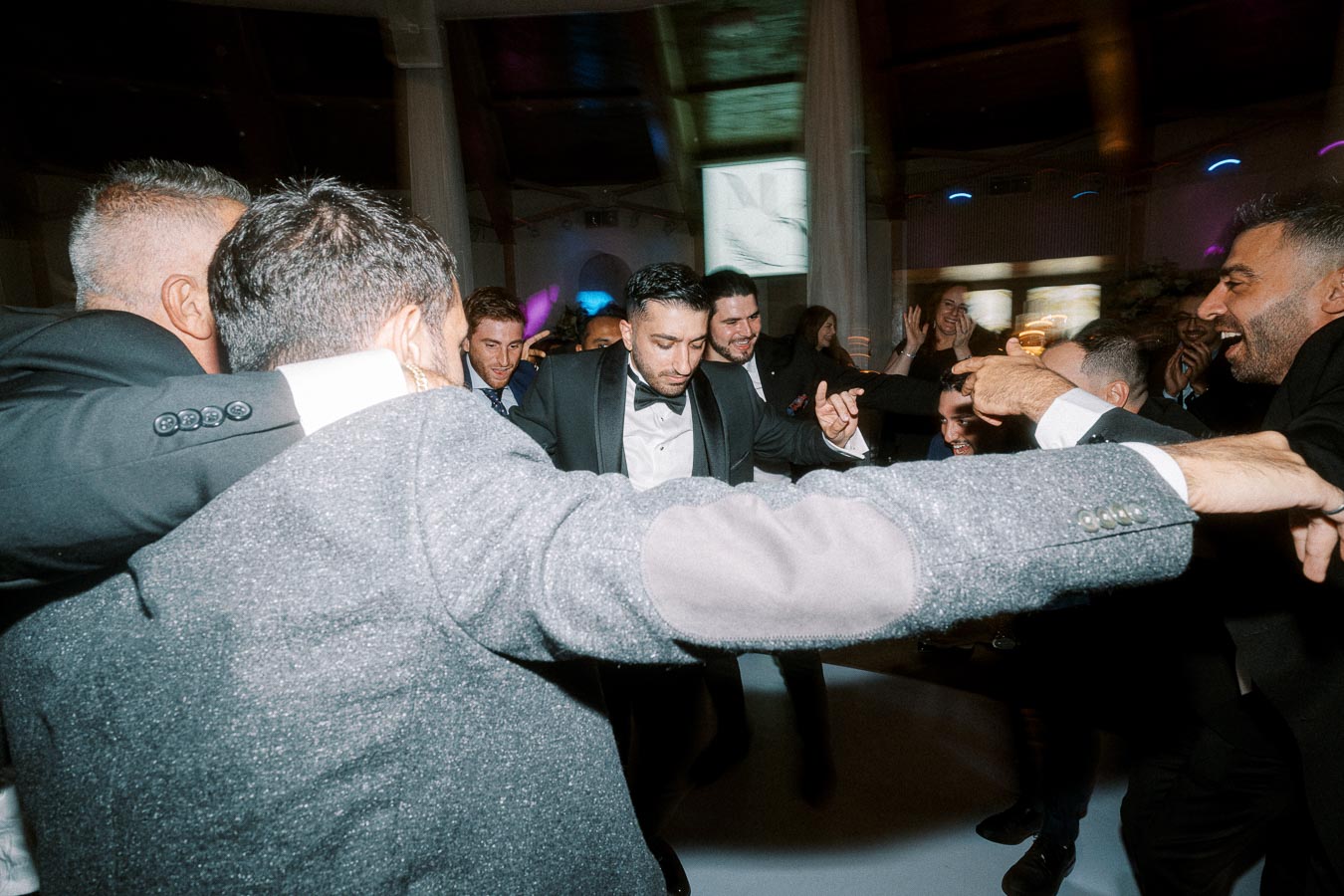 A group of well-dressed men enjoying a celebratory dance at an indoor event, with bright lights and a lively atmosphere.