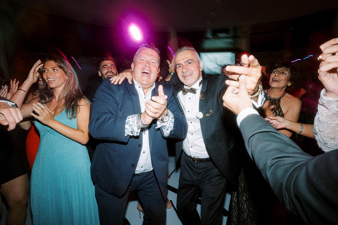 A group of people joyfully dancing and celebrating at a lively party, with colorful lights illuminating the scene. The men