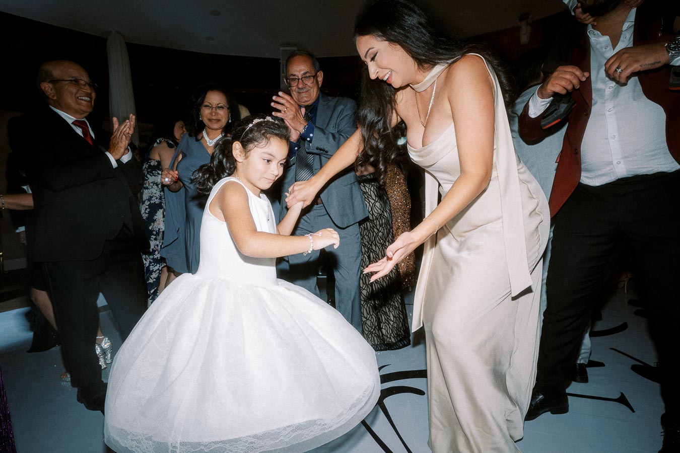 A joyful wedding celebration with guests dancing, featuring a woman in a beige dress twirling a young girl in a white dress