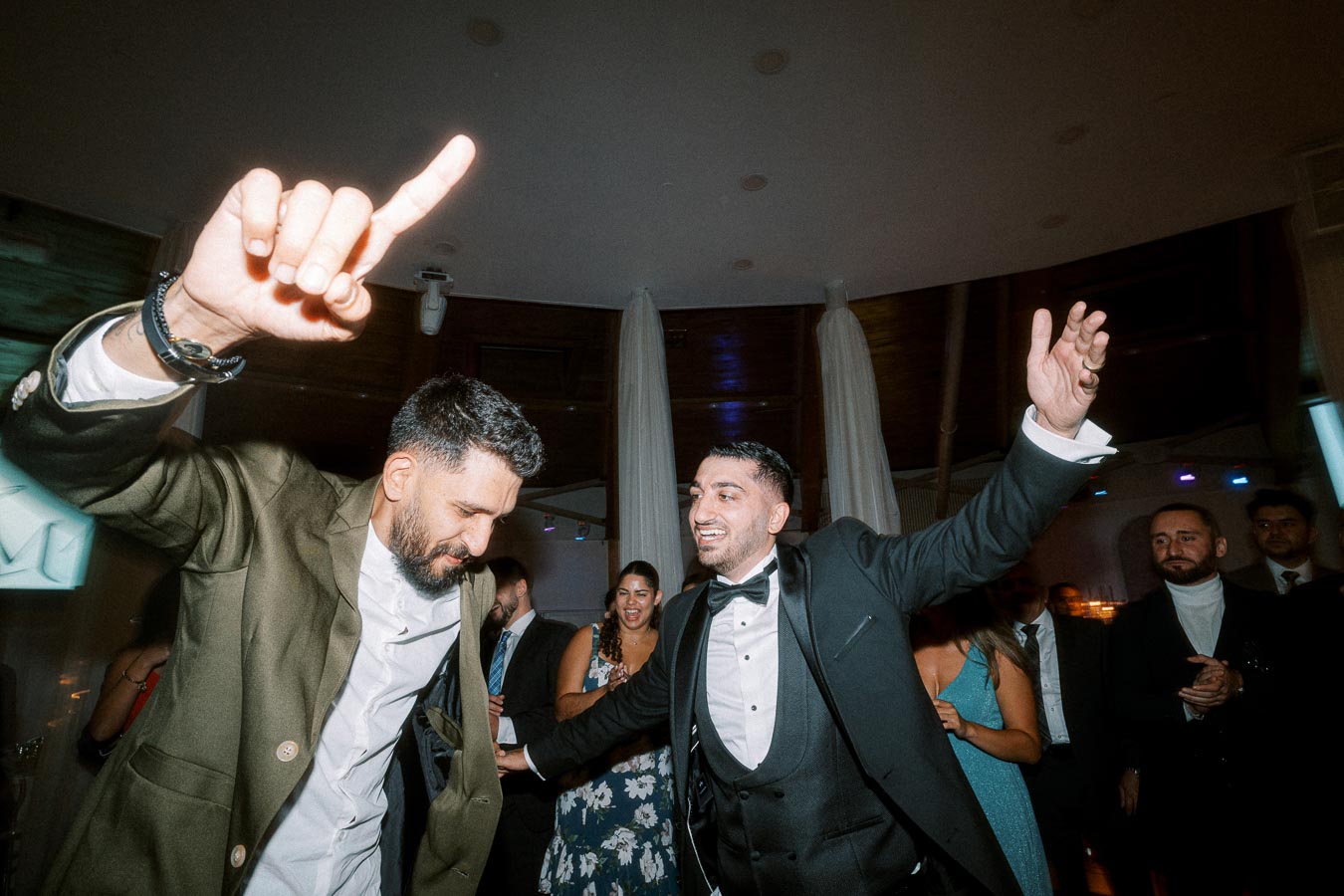 Two men joyfully dancing at a lively indoor celebration, surrounded by well-dressed guests, creating a festive and enjoyable