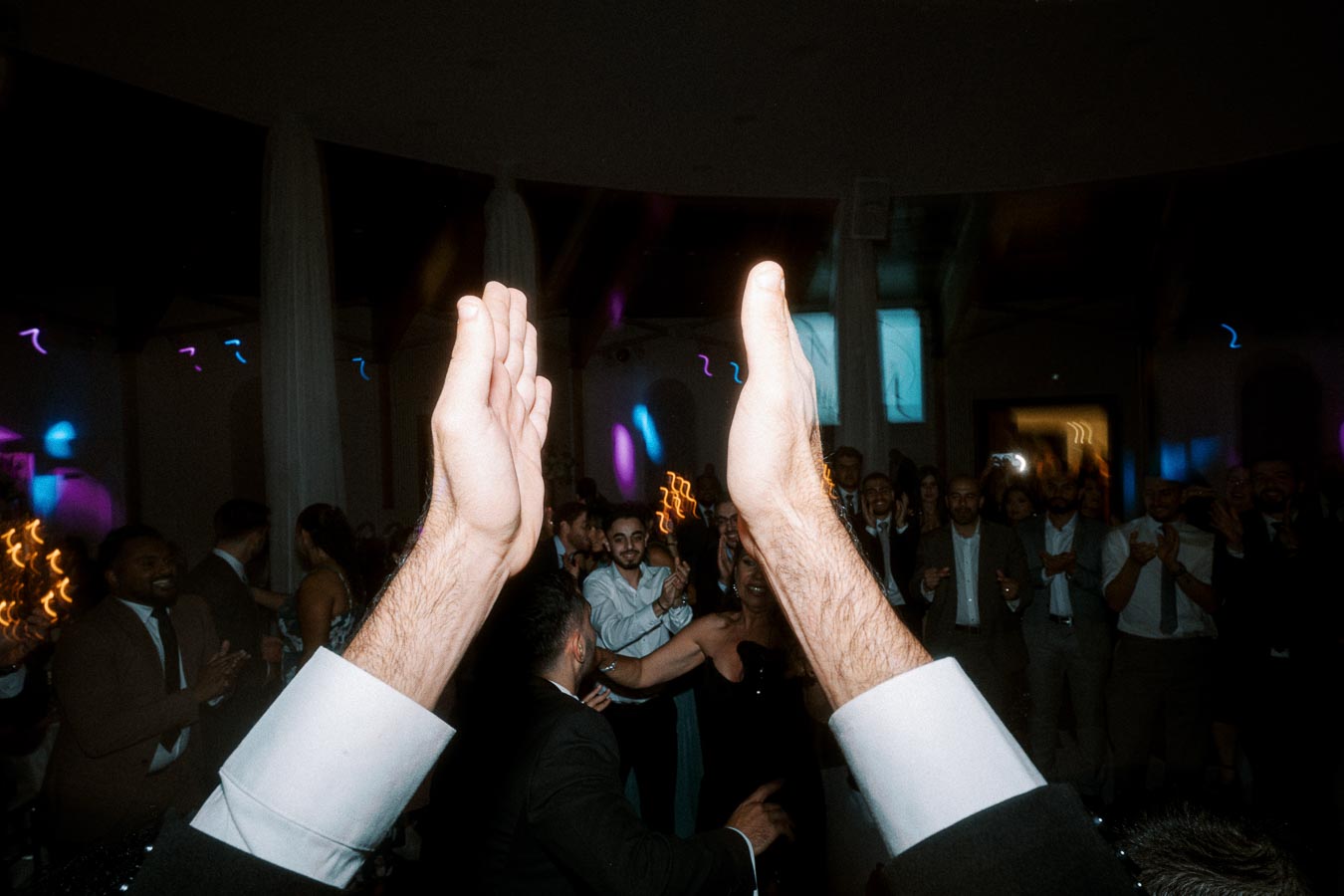 Clapping hands at a lively wedding reception with people dancing and celebrating against a backdrop of colorful lights.