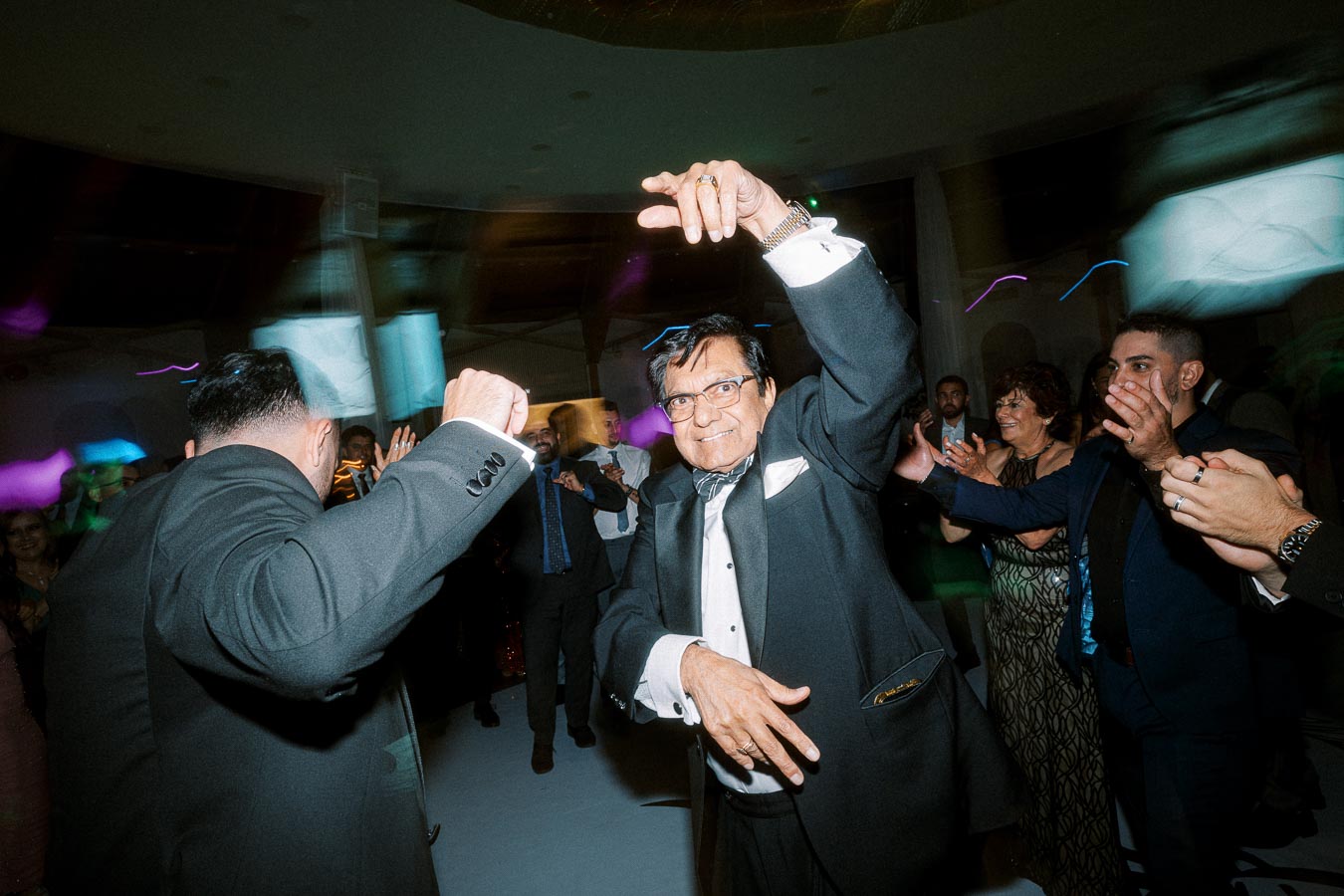 Elderly man in a tuxedo energetically dancing in a lively crowd at a formal indoor event.