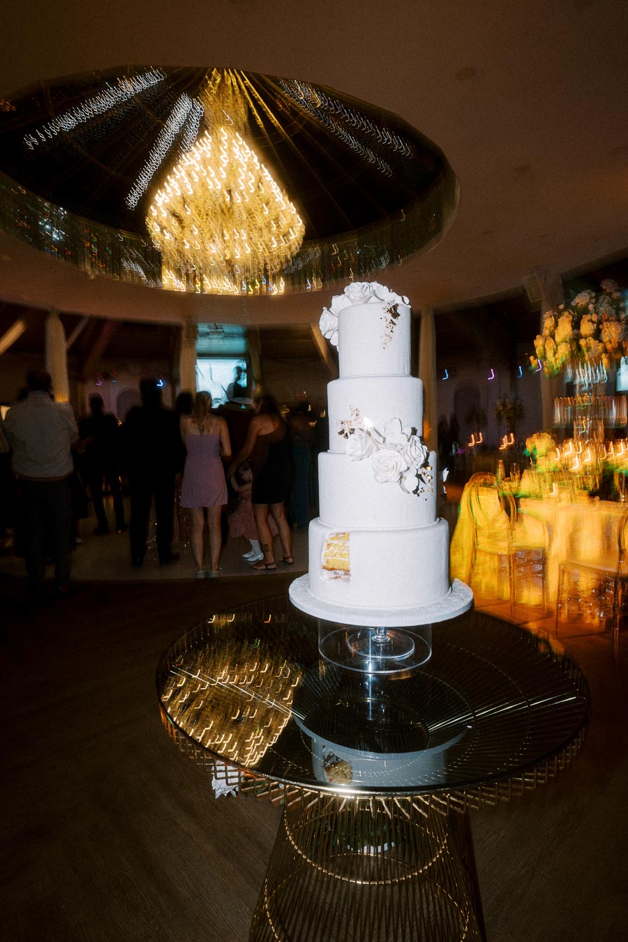 Four-tier white wedding cake with floral decorations, displayed on a reflective glass table in a dimly lit, elegant banquet