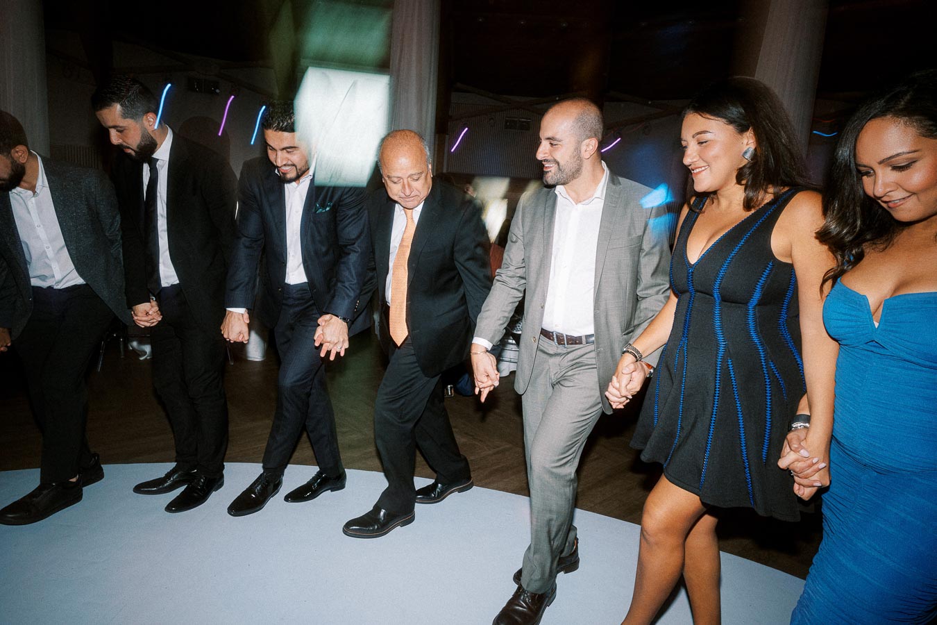 A group of people in formal attire joyfully dancing hand in hand at an indoor event, showcasing a lively and festive