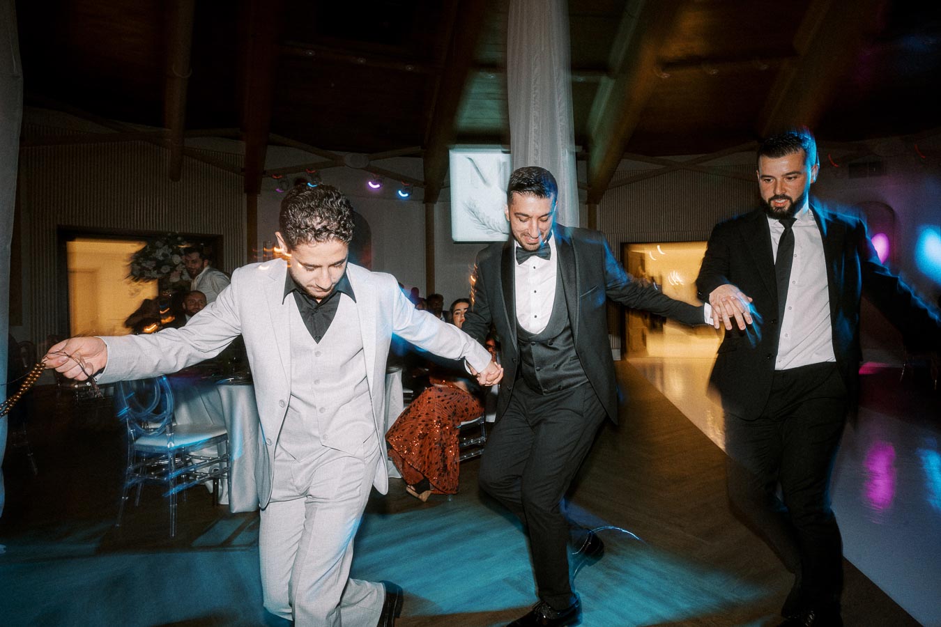 Three men in formal suits dancing joyfully at an indoor wedding reception, showcasing cultural and celebratory elements. The