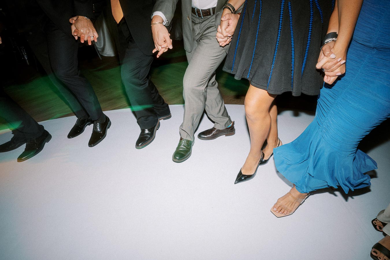 People holding hands and dancing in a circle at an indoor event, showcasing diverse footwear and elegant attire, including a