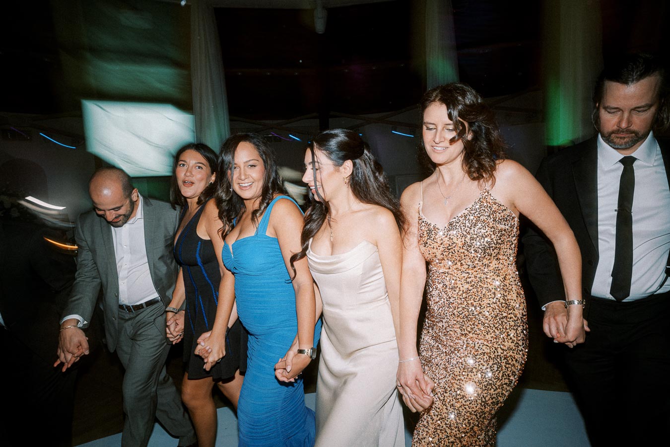 A group of elegantly dressed people holding hands and dancing at a lively event, showcasing formal attire and joyful