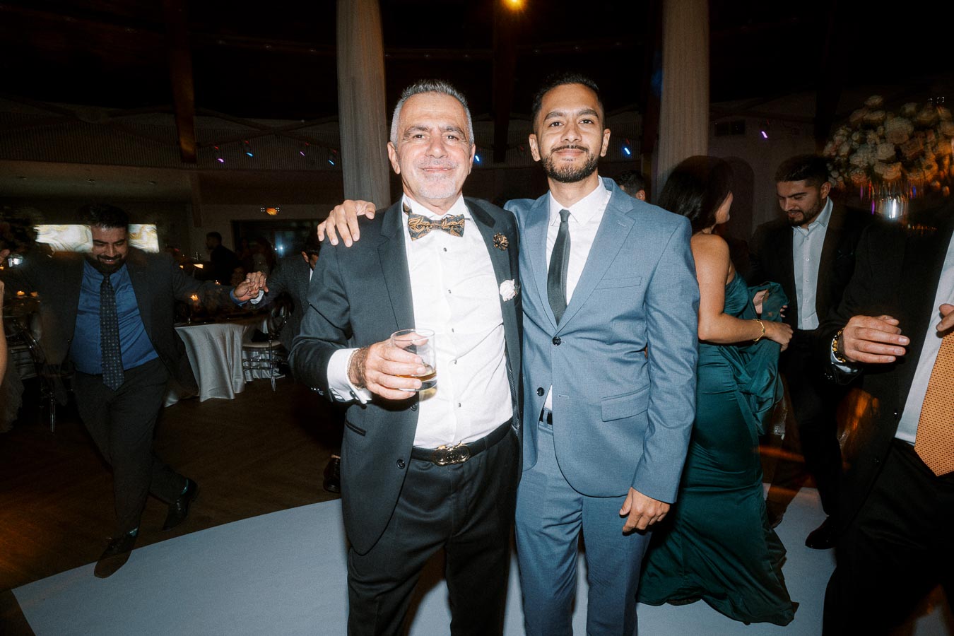 Two men in suits smiling and embracing at a lively indoor event, one holding a drink, surrounded by other guests in formal