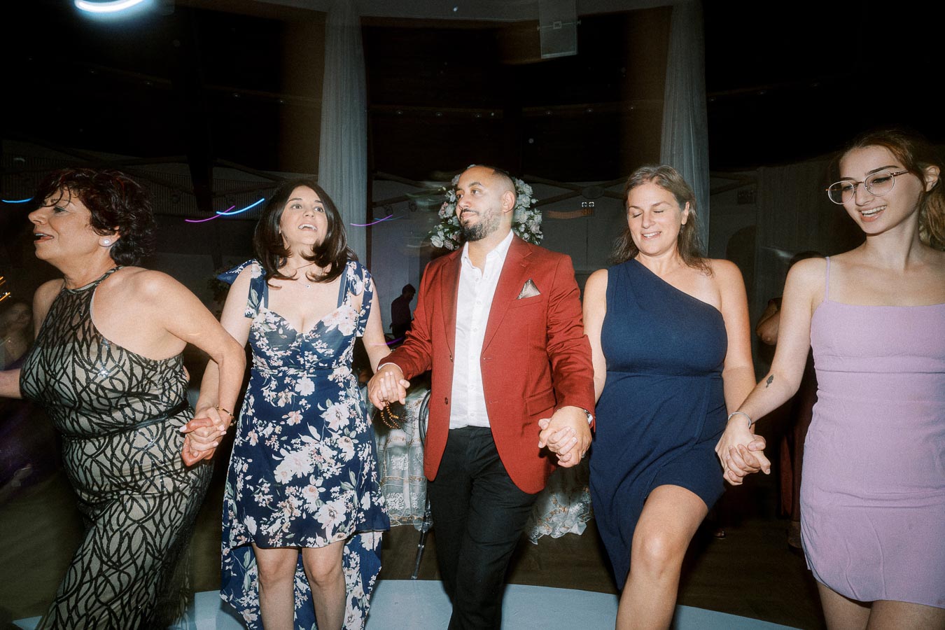 A group of people joyfully holding hands and dancing at a festive event, wearing a variety of elegant outfits including
