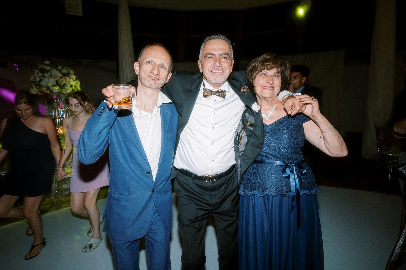 A group of people dressed formally at a wedding reception, standing together and smiling, with one person holding a drink.