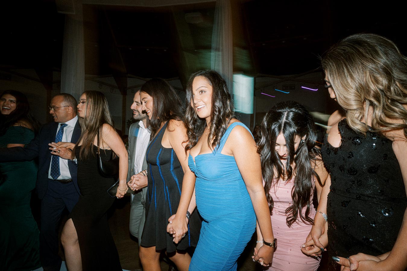 Group of people joyfully dancing and holding hands at an indoor event, with women in colorful dresses and a man in a suit,