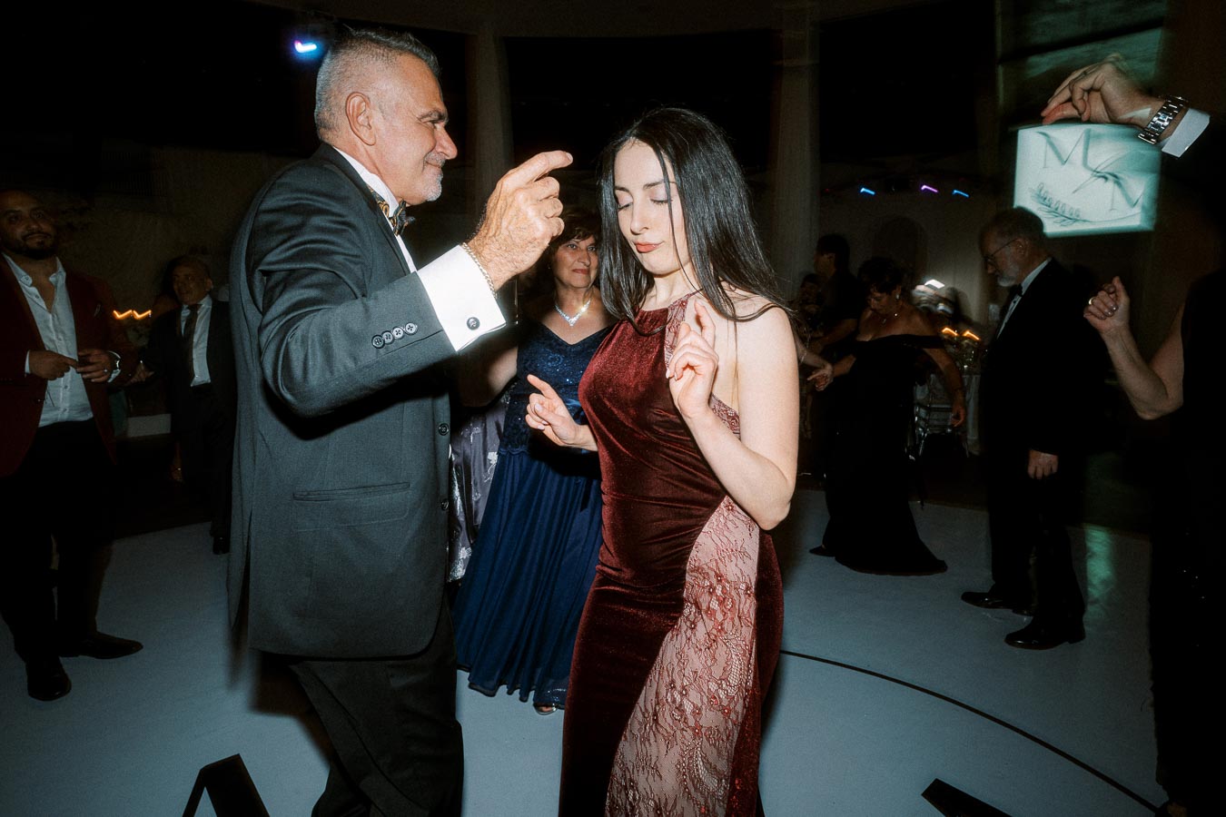 A lively indoor celebration with elegantly dressed guests dancing, featuring a man in a suit and a woman in a burgundy
