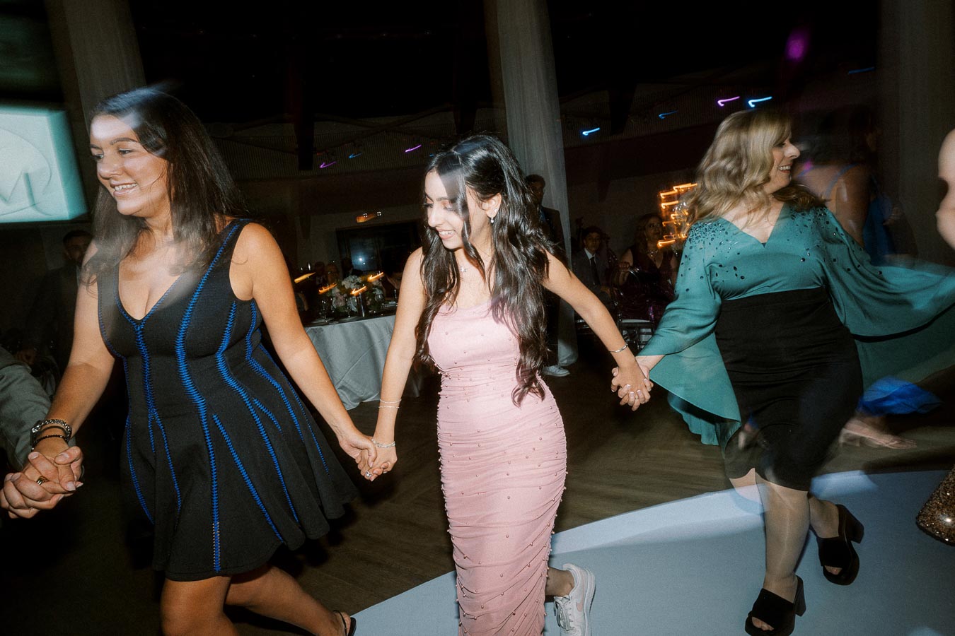 Women joyfully dancing at a celebration event, wearing elegant dresses, holding hands, and exhibiting happiness and energy