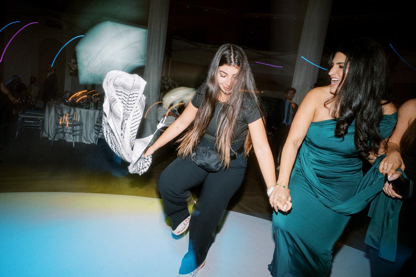 Two women joyfully dancing in an event setting, one wearing a black outfit and holding a patterned scarf, and the other in a
