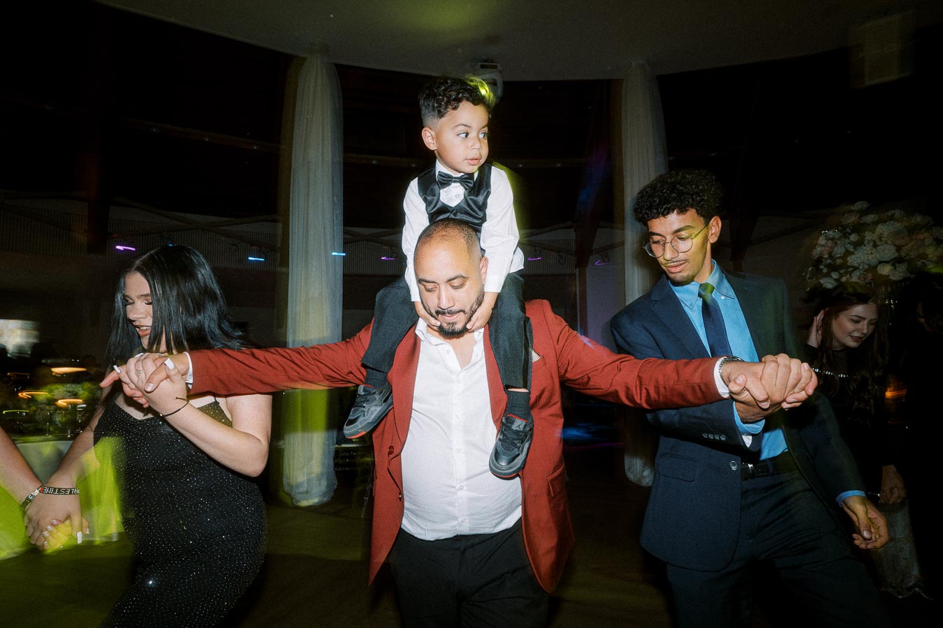 A joyful family dance at a wedding reception, featuring a man in a red jacket carrying a child on his shoulders while