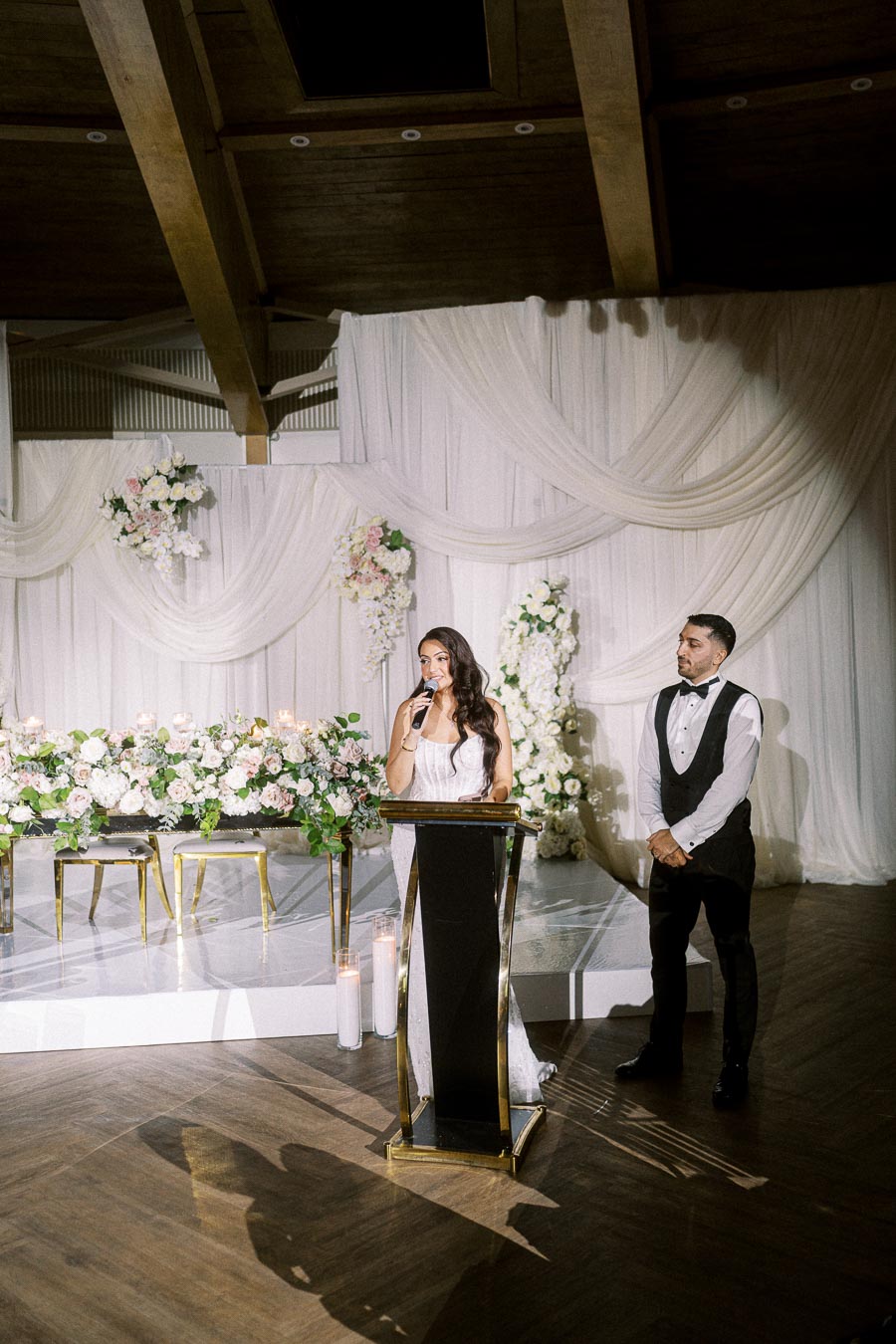 Bride giving a wedding speech at an elegant reception venue, beautifully decorated with white drapery and floral