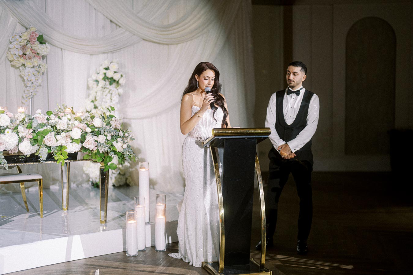 A woman in an elegant white gown speaks into a microphone at a podium during a formal wedding reception, with a man in a