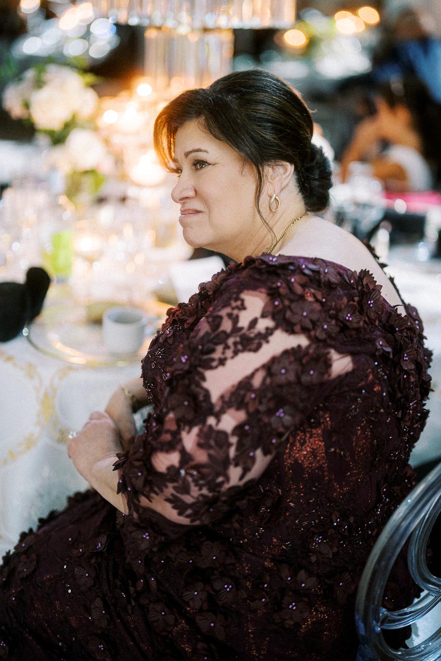 Elegant woman seated at a formal event wearing a detailed maroon lace dress, surrounded by a softly lit ambiance with