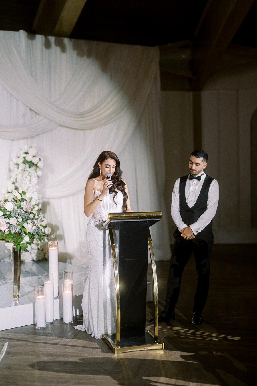 A woman in an elegant white dress gives a speech at a wedding reception podium, with a man in a black suit standing nearby,