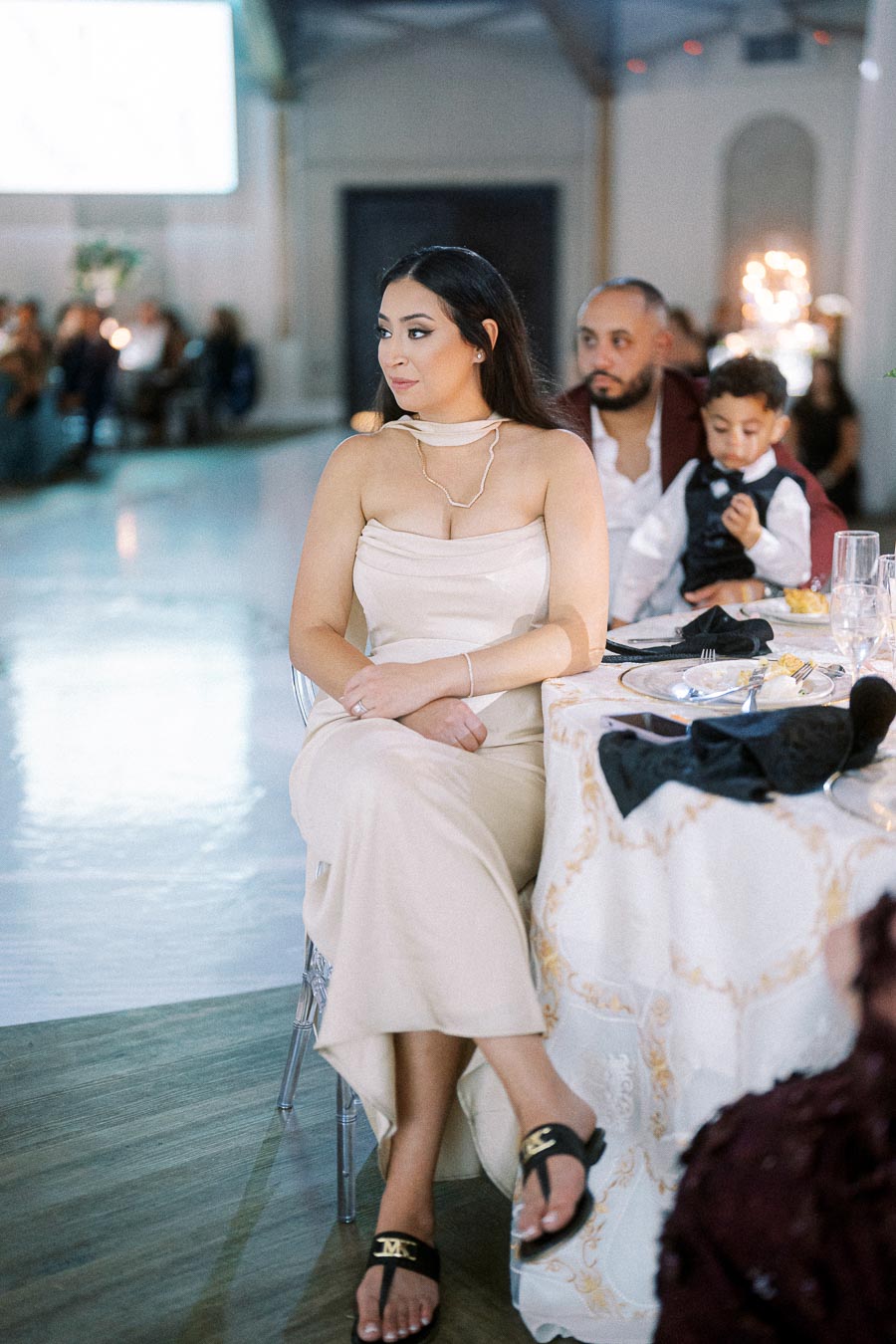 A woman in an elegant beige dress attending a formal event, seated beside a table set with plates and glasses. A man and a
