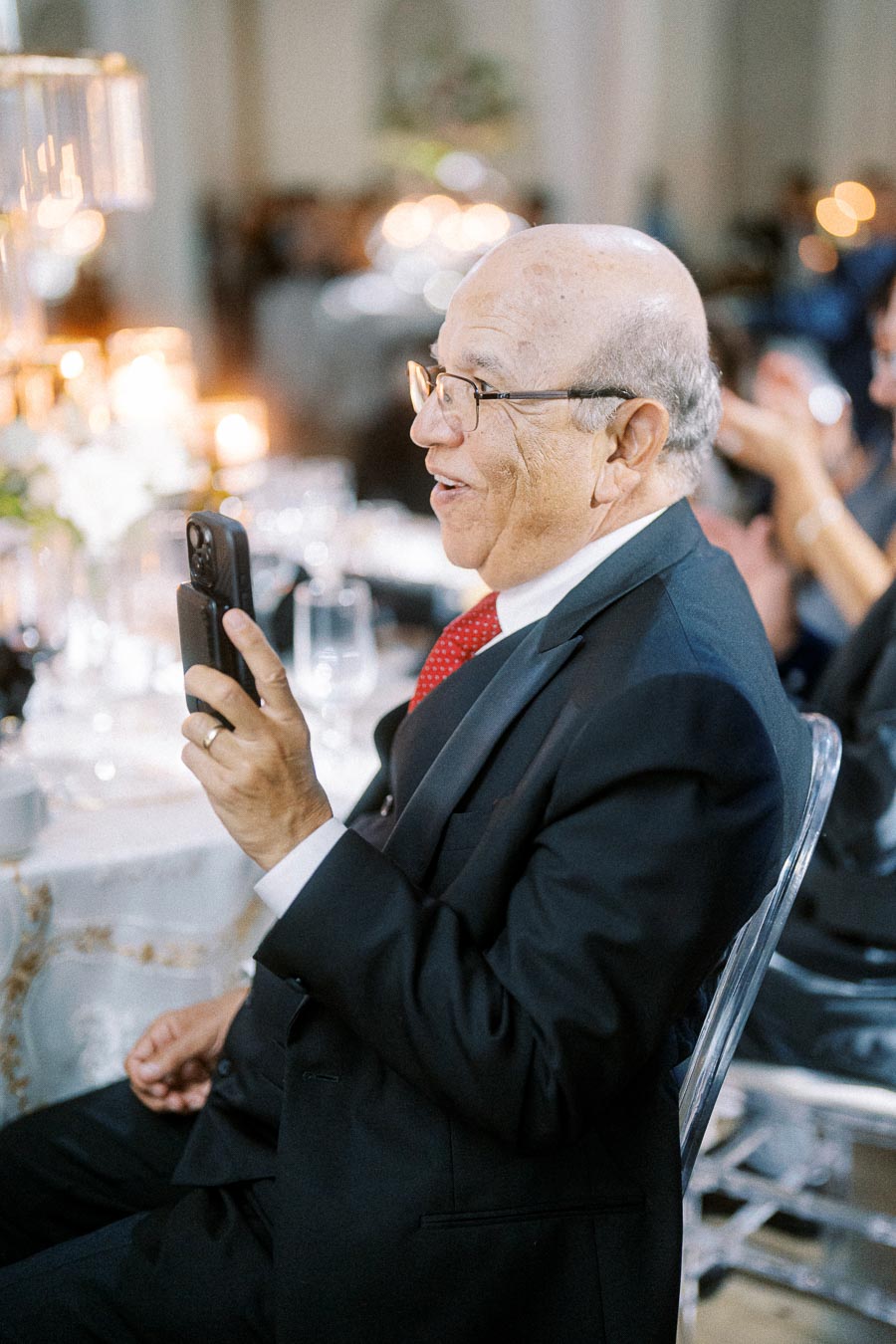 A smiling elderly man in a suit holding a smartphone at an elegant event, with tables set with candles and glassware in the