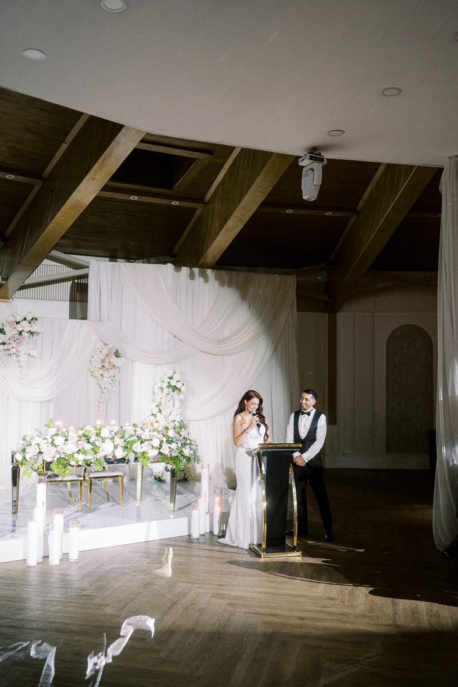 Elegant wedding scene with bride and groom standing at a podium, surrounded by white drapery and floral arrangements, in a