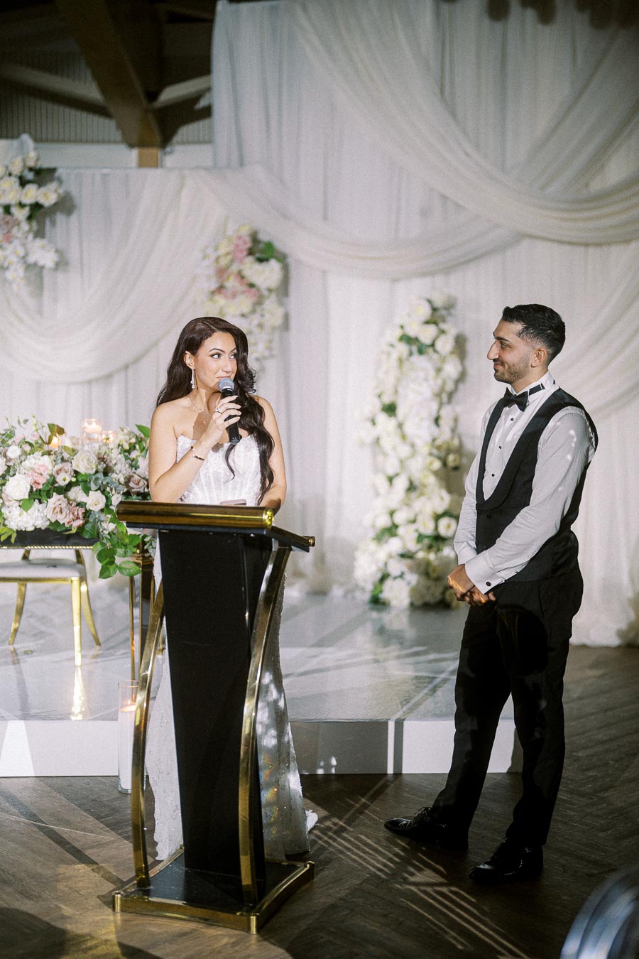 Elegant wedding reception with a woman in a white dress speaking at a podium, adorned with a floral arrangement, while a man