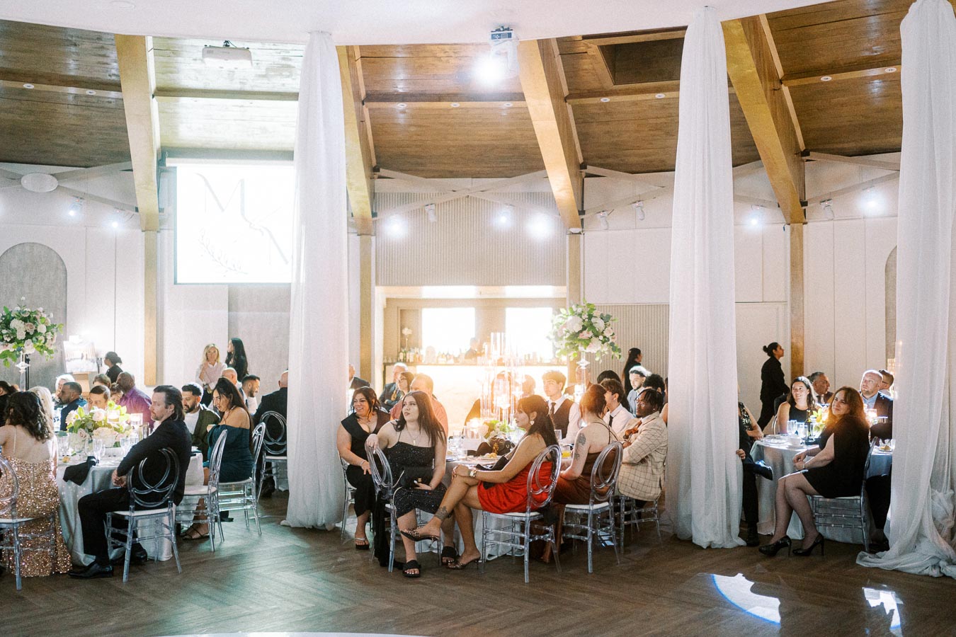 A group of elegantly dressed guests seated at round tables in a beautifully decorated reception hall with high ceilings and