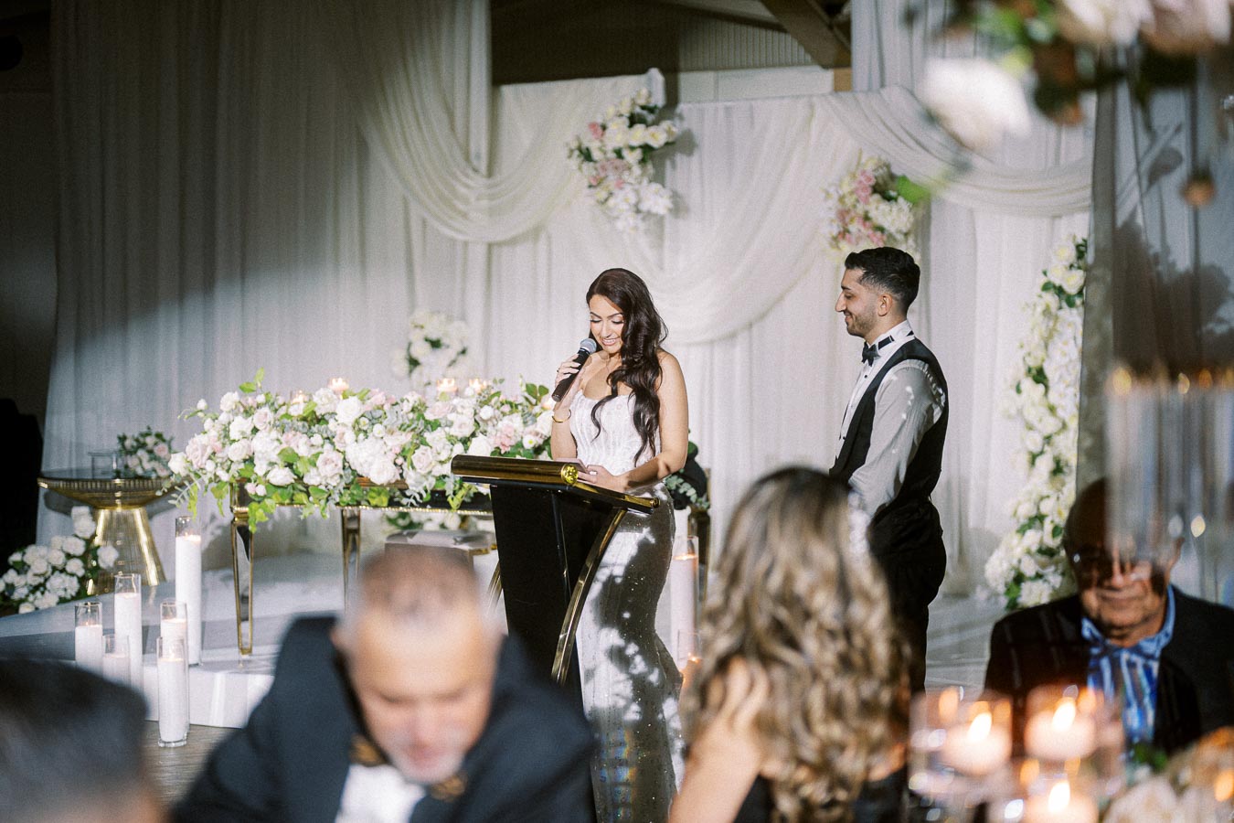 A woman in a formal gown gives a speech at a wedding reception, standing at a podium decorated with flower arrangements. A