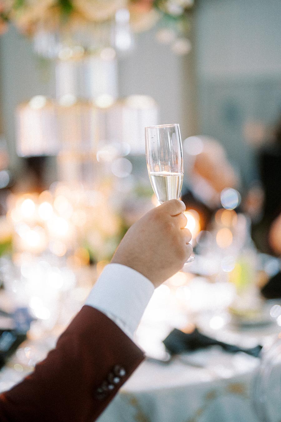 A hand holding a champagne flute in a formal celebration setting, with blurred background featuring soft lights and floral
