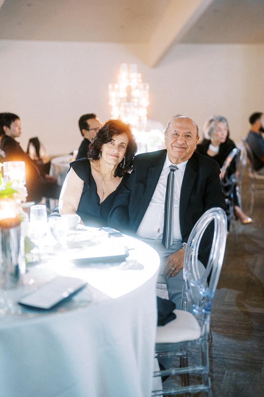 Elegant couple sitting at a beautifully set table during a formal event, with soft lighting and guests in the background