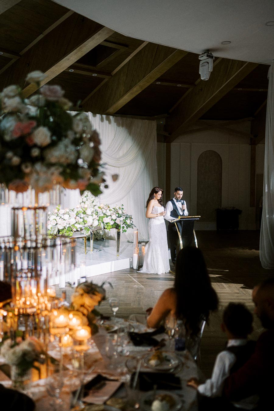 A bride and groom giving a speech at a wedding reception, with elegant floral arrangements and candlelit tables in an