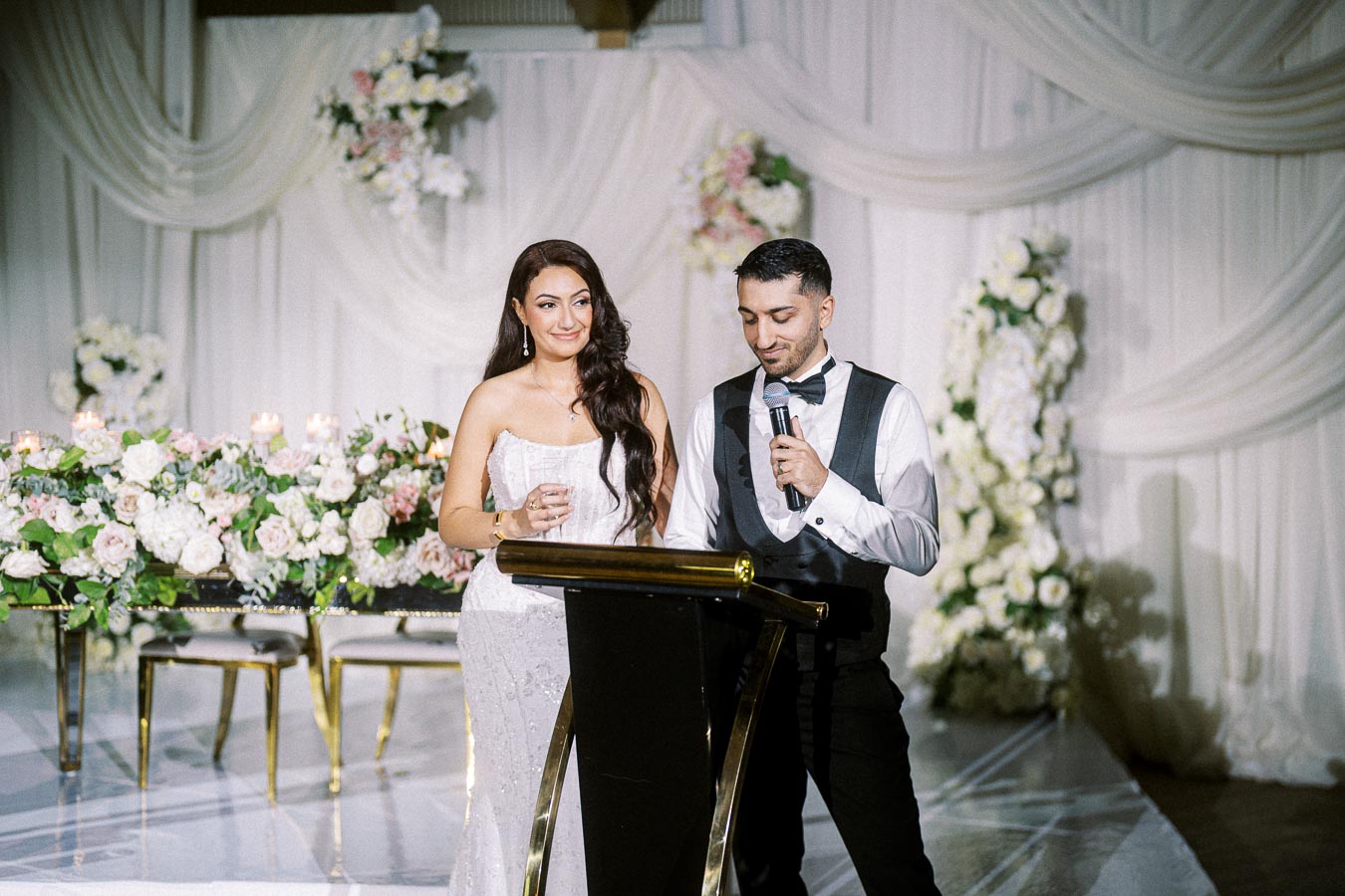 A couple giving a speech at a wedding reception with elegant floral decorations and draped white fabric in the background.