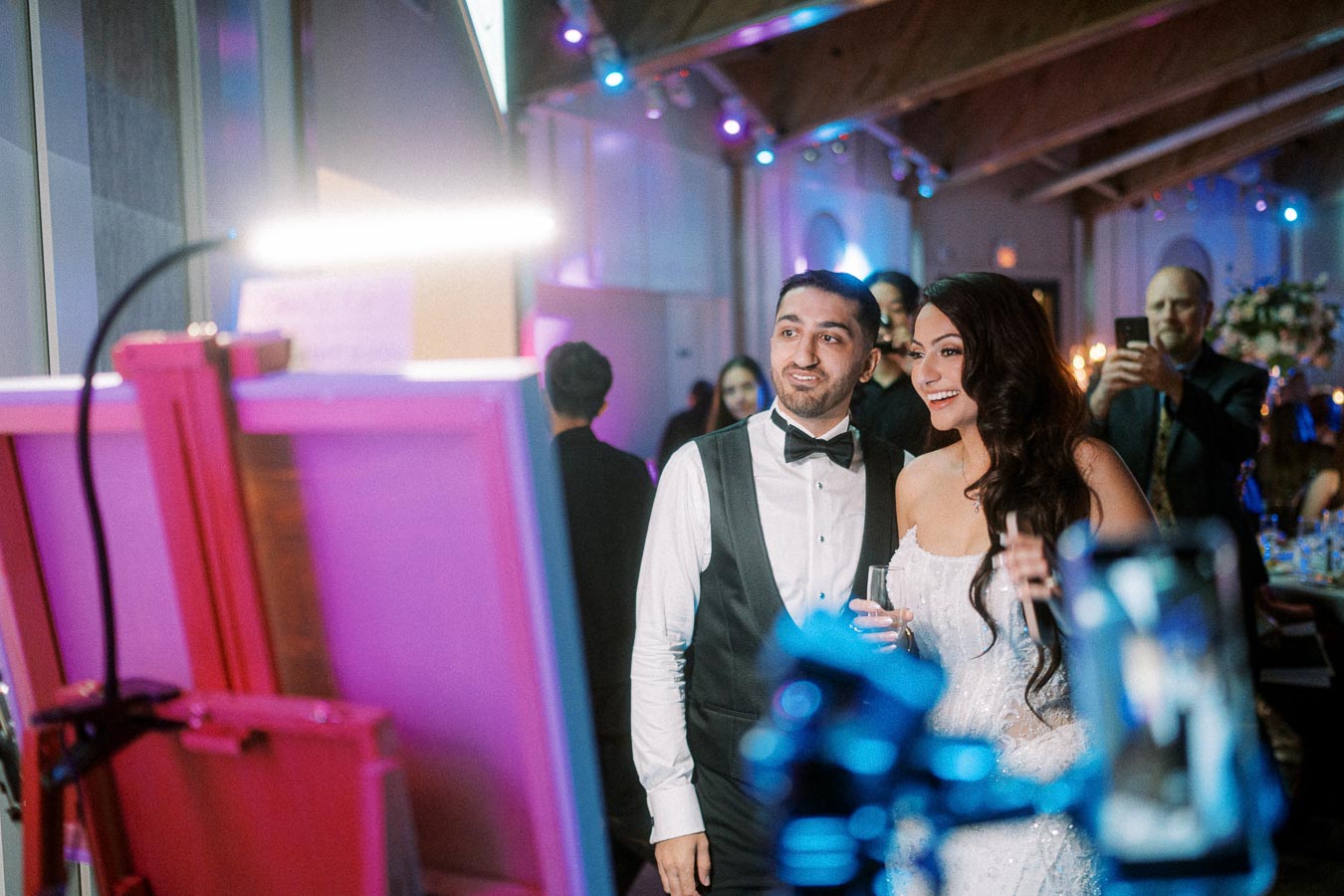 A couple in formal attire smiles while posing for a picture at a wedding reception, illuminated by vibrant lighting. The