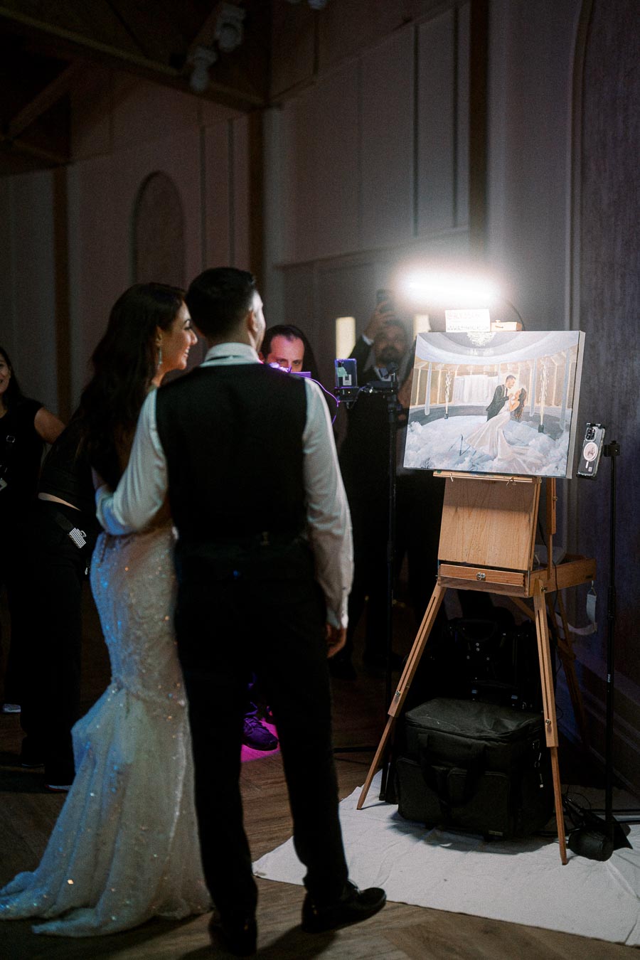 A couple admiring a live painting of themselves at a wedding reception, captured in a dimly lit venue with onlookers in the