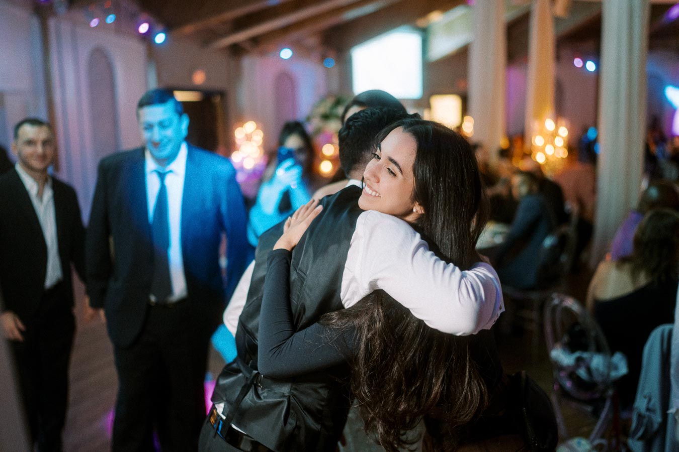 A joyful woman embracing a man at a lively event with warm ambient lighting, surrounded by people in formal attire.