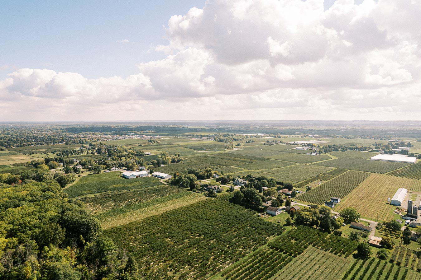 Aerial view of expansive farmland under a partly cloudy sky, showcasing lush green fields, scattered buildings, and distant horizon.