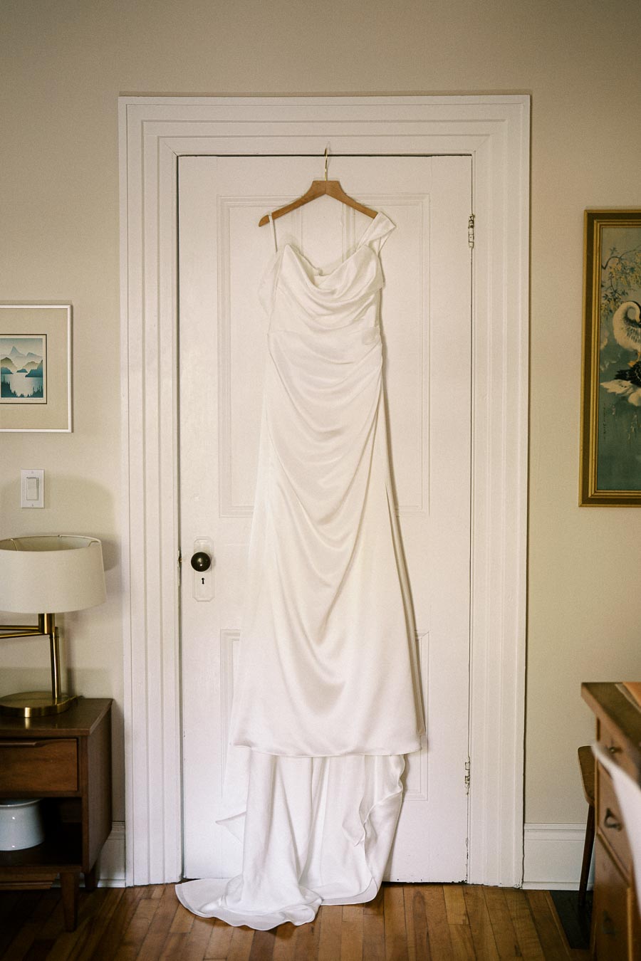 A simple and elegant white wedding dress hanging on a wooden hanger on a white door, set in a cozy room with framed artworks and a wooden table lamp.